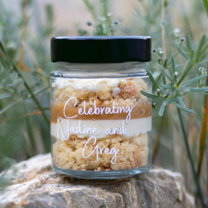 layers of cake and frosting in a jar for a birthday party with custom label for wedding guests saying celebrating nadine and greg